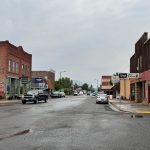 Elgin OR 28 A small town street with wet pavement, lined with brick and wooden buildings, parked cars, and a few vehicles driving. Overcast sky with mountains faintly visible in the background. Elgin, OR