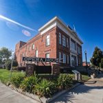 Elgin OR 10 A historic red-brick building with white trim stands under a clear blue sky in Elgin, OR. A sign in front reads Opera House, surrounded by green plants and a sidewalk leading up to the entrance.