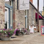 Elgin OR 27 A stone building with flower planters and an OPEN sign outside a hair salon called Hairitage Salon. The sidewalk is wet, and there are patriotic decorations on the planters.