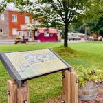 Elgin OR 24 A park scene with an informational sign in the foreground, next to a barrel planter with flowers. In the background, there are historic brick buildings, green grass, and leafy trees on a cloudy day.