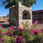 Elgin OR 22 A stone wishing well stands in a sunny Elgin courtyard, surrounded by blooming purple and pink flowers, with a tree and buildings in the background.