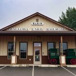 Elgin OR 19 A small train depot building labeled Elgin Wallowa Union Railroad with a covered porch, green luggage carts, and a parked train visible on the left. Parking spaces in front are marked for conductor and depot use.