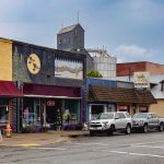 Elgin OR 26 A small-town main street with parked cars, colorful storefronts, and murals on building walls under a cloudy sky. A grain elevator and more buildings are visible in the background.
