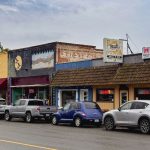 Elgin OR 30 A row of small shops and restaurants with colorful facades lines a street. Several cars are parked in front. The sky is overcast, and signs for businesses and neon OPEN lights are visible.