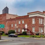 Elgin OR 12 A historic red brick building labeled “Opera House” and “City Hall” sits on a corner with a street sign and greenery. The sky is overcast and the street appears quiet with no visible pedestrians.