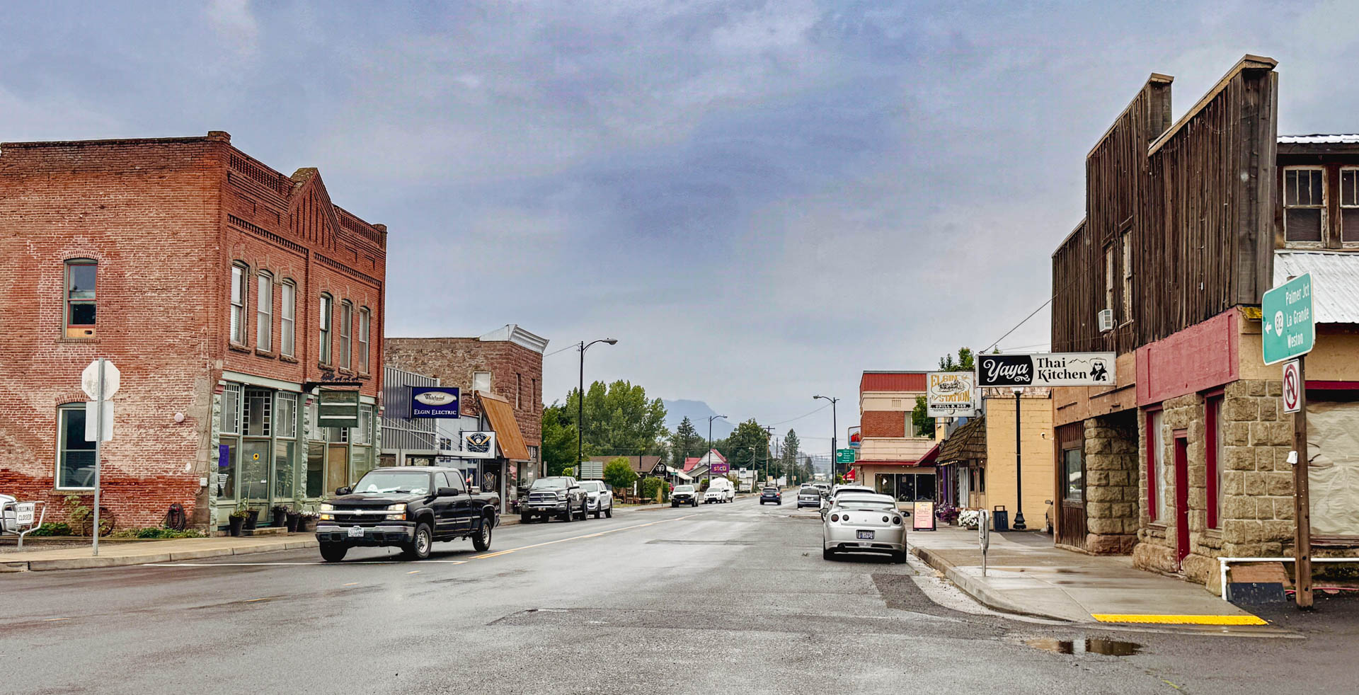 Elgin OR 2 A small-town main street with brick and stone buildings, parked cars, storefront signs, and a cloudy sky. A mountain is faintly visible in the distance at the end of the street.