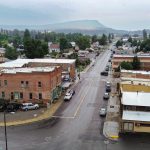 Elgin OR 21 Aerial view of a small town with a main street lined by brick and wooden buildings, cars parked along the road, and houses surrounded by trees, with hills and cloudy sky in the background.