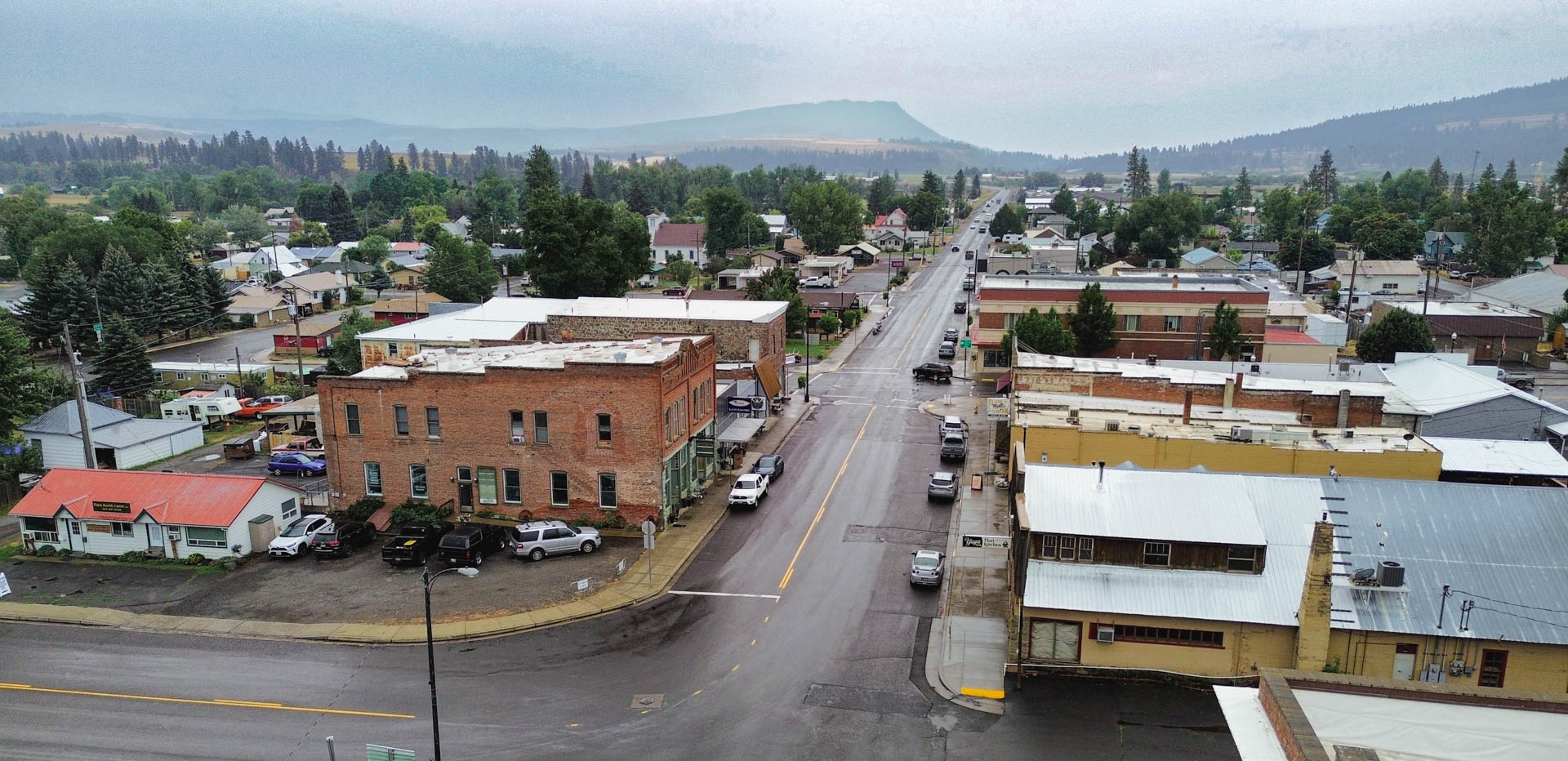 Elgin OR 5 Aerial view of a small town with a main street lined by brick and wooden buildings, cars parked along the road, and houses surrounded by trees, with hills and cloudy sky in the background.