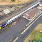 Elgin OR 16 Aerial view of a small train station with a long passenger train stopped at the platform, surrounded by empty parking lots, tracks, and grassy areas.