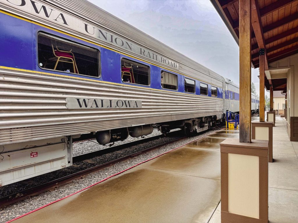 Elgin OR 14 A silver and blue passenger train labeled WALLOWA UNION RAILROAD is stopped at a wet, covered platform on a rainy day. Empty chairs are visible through the windows of the train car.