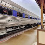 Elgin OR 18 A silver and blue passenger train labeled WALLOWA UNION RAILROAD is stopped at a wet, covered platform on a rainy day. Empty chairs are visible through the windows of the train car.