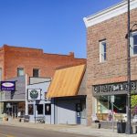 Elgin OR 32 A small-town main street in Elgin, OR, with historic brick and stone buildings—including shops and the City Drug pharmacy—under a clear blue sky. Tables and chairs sit outside some storefronts.