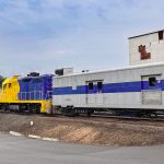 Elgin OR 17 A yellow and blue locomotive pulls a silver and blue passenger car past industrial buildings and silos under a cloudy sky.