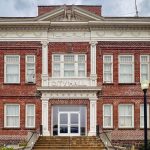 Elgin OR 9 A historic two-story red brick building with white columns and trim, labeled CITY HALL above the entrance, flanked by two lamp posts and steps leading to double glass doors.