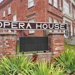 Elgin OR 11 A large “Opera House” sign is mounted between two brick pillars, with a historic red brick building in the background and green plants in the foreground.
