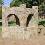 Elgin OR 31 A stone structure with two arched openings stands on a paved area in Elgin's grassy park, with benches nearby and trees, a fence, and buildings in the background.