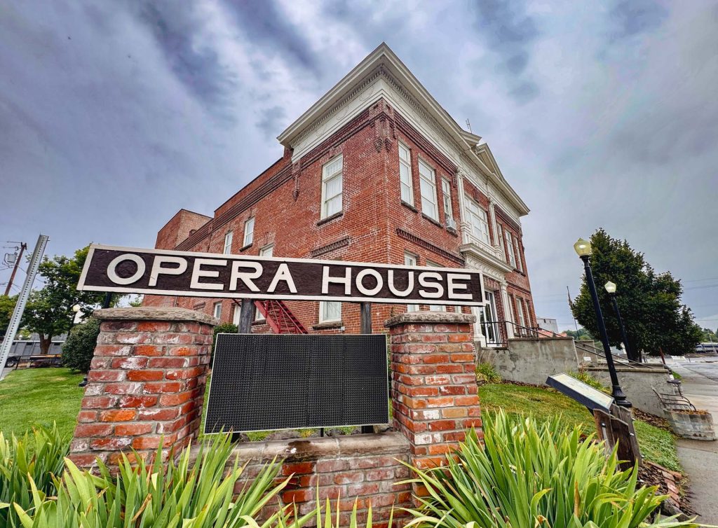 Elgin OR 7 A large brick building with white trim stands behind a sign that reads Opera House in bold white letters. The sign is mounted on two brick pillars, surrounded by green plants under a cloudy sky.