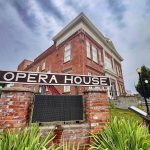 Elgin OR 13 A large brick building with white trim stands behind a sign that reads Opera House in bold white letters. The sign is mounted on two brick pillars, surrounded by green plants under a cloudy sky.