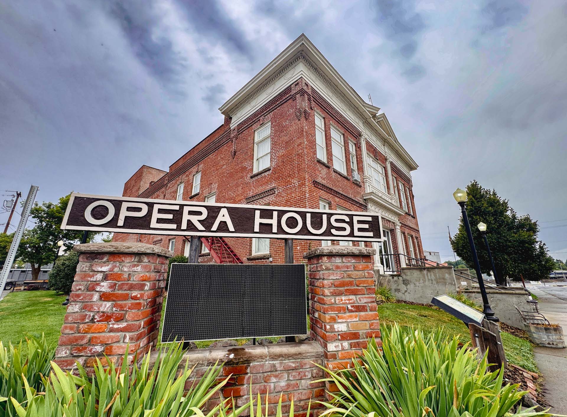Elgin OR 3 A large brick building with white trim stands behind a sign that reads Opera House in bold white letters. The sign is mounted on two brick pillars, surrounded by green plants under a cloudy sky.
