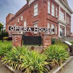 Elgin OR 8 A historic red-brick building with white trim, labeled “Opera House” on a large sign in front, surrounded by green plants and a wet sidewalk under a cloudy sky.