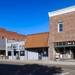 Elgin OR 25 Small-town main street in Elgin, OR, with historic brick buildings—including City Drug pharmacy, a bank, and a coffee shop—under a clear blue sky. The peaceful street is quiet, with no vehicles or pedestrians in sight.