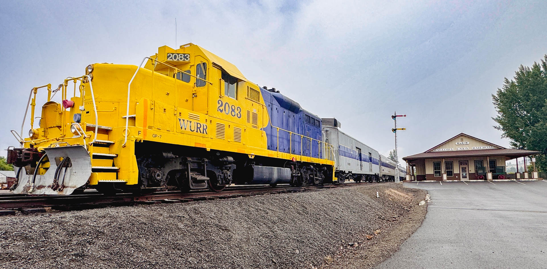 Elgin OR 4 A bright yellow and blue train labeled 2088 on the front sits on railroad tracks near a small train station building with Yreka Western Railroad sign, under a cloudy sky.